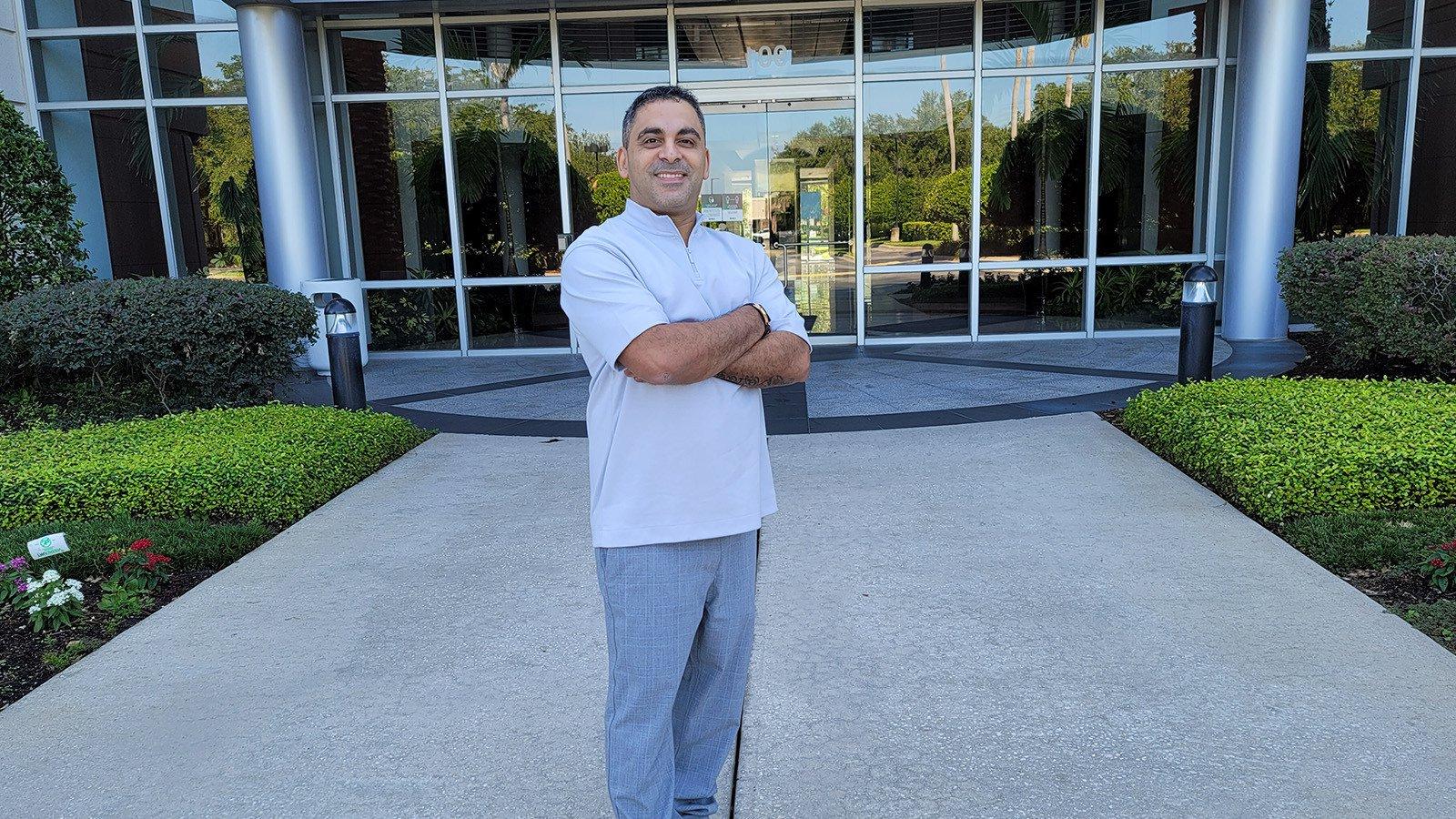 Brian Alvarado stands smiling with his arms crossed in front of the Deloitte building. He wears gray slacks and a blue shirt.