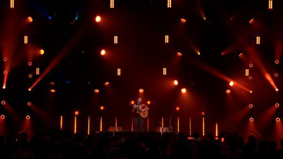 Trivium乐队主唱Matt Heafy站在灯光昏暗的舞台上，在一大群人面前弹着原声吉他.