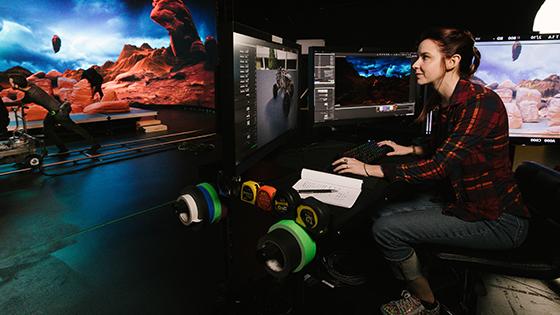 一个女人坐在桌子前，面前是几台显示器. 她在一个虚拟的制作工作室里，这个工作室被设计得看起来和火星很像.